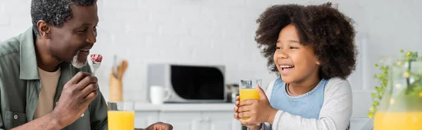 Happy african american girl holding glass of orange juice while having breakfast with grandfather, banner — Stock Photo
