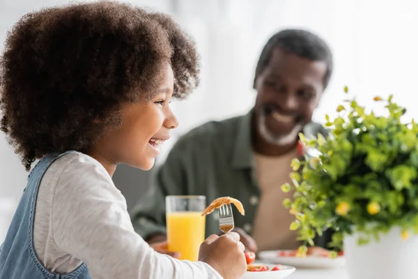 Feliz niño afroamericano feliz sosteniendo tenedor con panqueque mientras desayuna con el abuelo - foto de stock