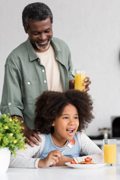 Abuelo afroamericano feliz sosteniendo un vaso de jugo de naranja y de pie detrás de la nieta durante el desayuno - foto de stock