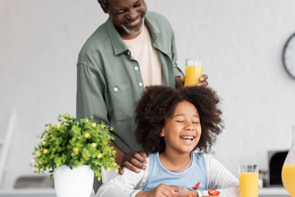 Feliz avô afro-americano segurando vidro de suco de laranja e de pé atrás criança alegre durante o café da manhã — Fotografia de Stock
