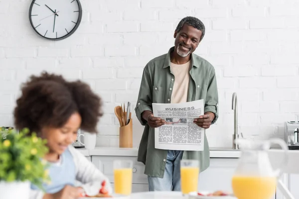 Feliz afroamericano abuelo leyendo periódico mientras nieta desayunando - foto de stock
