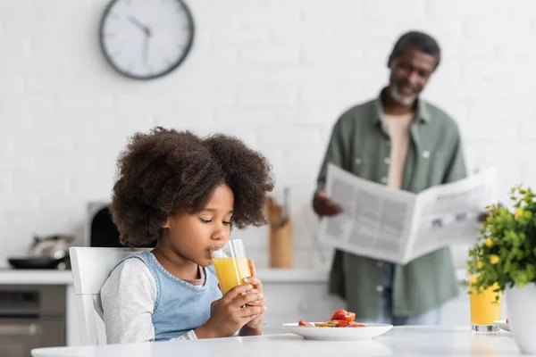 Encaracolado menina americana africana beber suco de laranja, enquanto avô ler jornal na cozinha — Fotografia de Stock