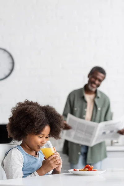 Happy african american kid drinking orange juice near grandfather reading newspaper on blurred background — Stock Photo