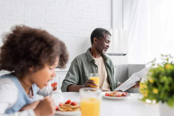 Happy african american grandparent reading newspaper while granddaughter having breakfast in kitchen — Stock Photo