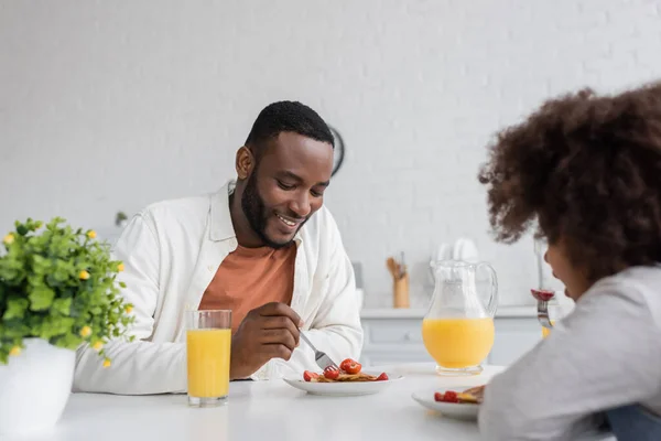 Hombre afroamericano feliz desayunando con su hija rizada en la cocina - foto de stock