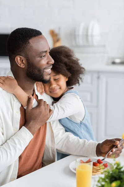 Rizado africano americano chica sonriendo mientras abrazando feliz padre durante el desayuno - foto de stock