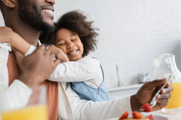 Lockiges afrikanisch-amerikanisches Mädchen lächelt, während es seinen fröhlichen Vater beim Frühstück umarmt — Stockfoto