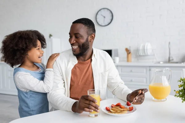 Menina americana africana feliz olhando para o pai alegre tomando café da manhã na cozinha — Fotografia de Stock
