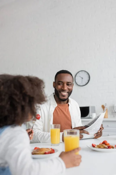 Heureux homme afro-américain tenant journal et regardant fille frisée prendre le petit déjeuner au premier plan flou — Photo de stock