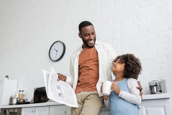 Happy african american man holding newspaper and looking at daughter with cup of tea — Stock Photo