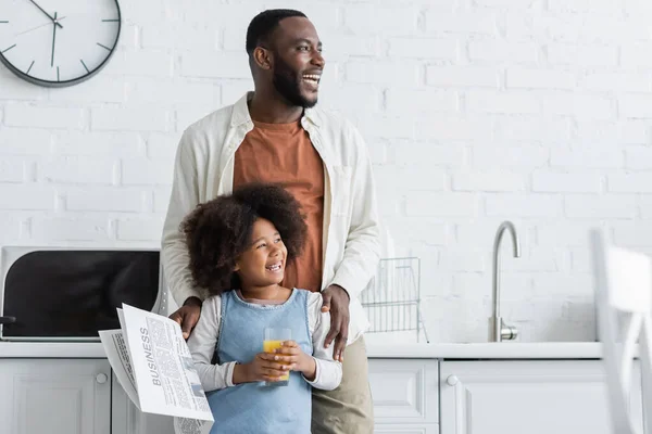 Happy african american man holding newspaper and hugging cheerful daughter with glass of orange juice — Stock Photo