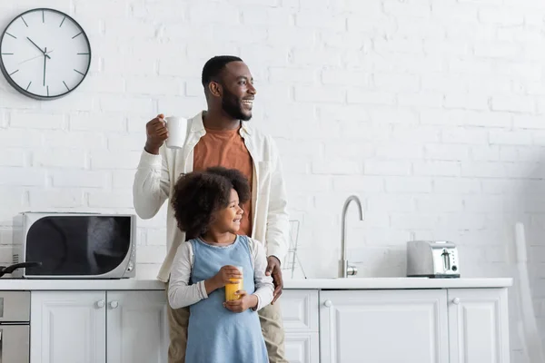 Счастливый африканский американец держит чашку кофе и обнимает веселую дочь стаканом апельсинового сока — стоковое фото