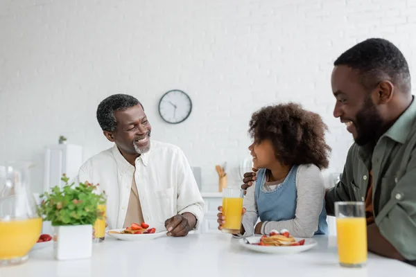 Счастливый мужчина среднего возраста смотрит на африканскую внучку рядом с сыном во время семейного завтрака — стоковое фото