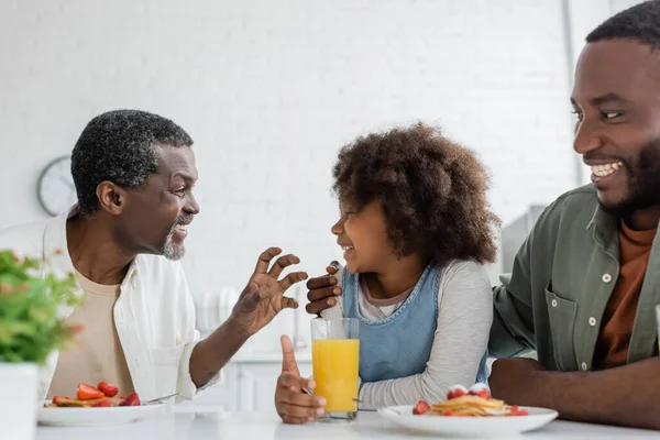 Lächelnder Mann mittleren Alters, der beim Familienfrühstück seine Enkelin in der Nähe eines glücklichen Sohnes ansieht — Stockfoto