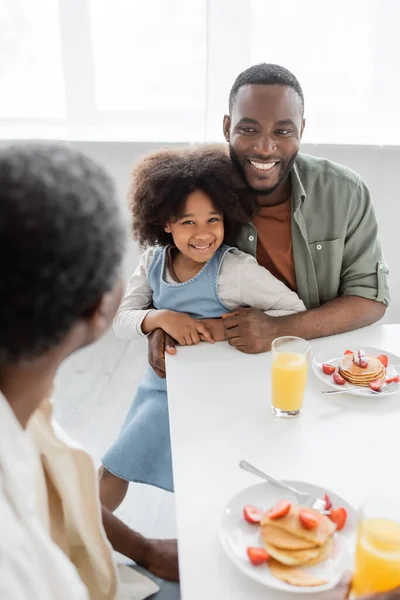 Счастливый африканский американец обнимает кудрявую дочь и смотрит на отца во время семейного завтрака — стоковое фото