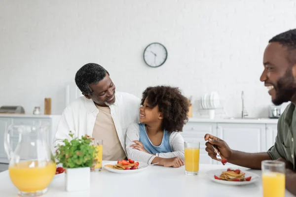 Щасливий афроамериканський чоловік обіймає кучеряву онуку біля веселого сина під час сімейного сніданку — стокове фото