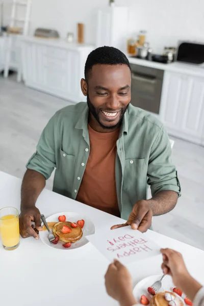 African american girl giving greeting card with happy fathers day lettering to cheerful dad during breakfast — Stock Photo