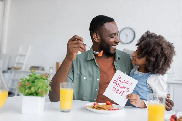 Cheerful african american girl holding greeting card with happy fathers day lettering near smiling dad during breakfast — Stock Photo