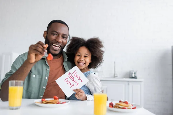 Curly african american girl holding greeting card with happy fathers day lettering near cheerful dad during breakfast — Stock Photo