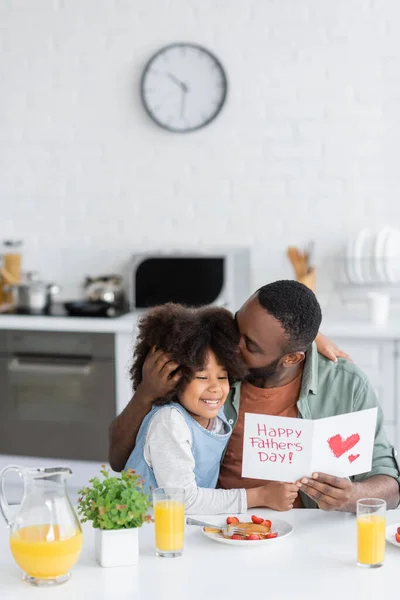 African american man holding greeting card with happy fathers day lettering while kissing head of curly daughter — Stock Photo