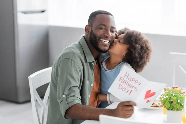 Curly african american girl kissing cheek of happy father with greeting card on fathers day — Stock Photo