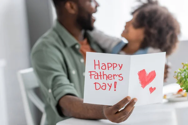 Africano americano uomo in possesso di biglietto di auguri con felice padri giorno lettering vicino riccio figlia su sfondo sfocato — Foto stock