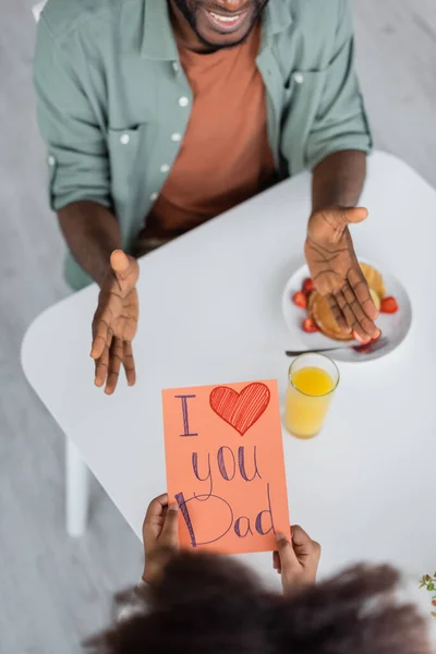 Top view of african american kid holding greeting card with i love you dad lettering near cheerful man on fathers day — Stock Photo
