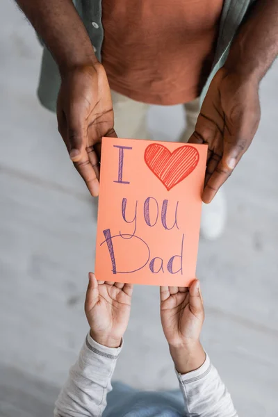 Vista superior del niño afroamericano recortado sosteniendo la tarjeta de felicitación con te amo papá letras cerca del hombre en el día de los padres - foto de stock