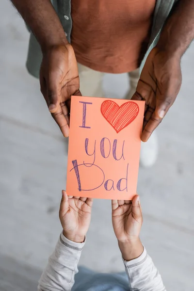 Top view of cropped african american kid holding greeting card with i love you dad lettering near man on fathers day — Stock Photo