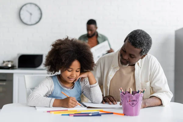 Happy african american girl with curly hair drawing near bearded grandfather at home — Stock Photo