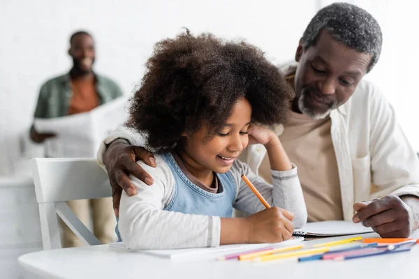 Bearded african american grandpa hugging happy granddaughter drawing on paper with color pencil — Stock Photo