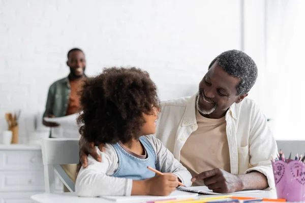 Bearded african american grandpa hugging happy kid drawing on paper with color pencil — Stock Photo
