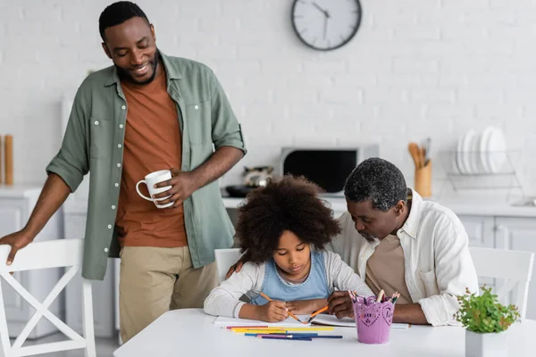 Felice uomo africano americano in possesso di tazza e guardando figlia disegno su carta vicino a suo padre — Foto stock