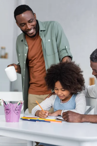 Felice uomo africano americano in possesso di tazza e in piedi vicino figlia disegno su carta vicino nonno — Foto stock
