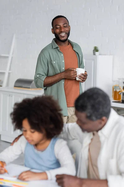 Heureux homme afro-américain tenant tasse et regardant fille dessin près de grand-père sur le premier plan flou — Photo de stock