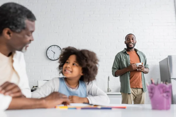 Hombre afroamericano feliz sosteniendo la taza y mirando a su hija y su padre en primer plano borrosa - foto de stock