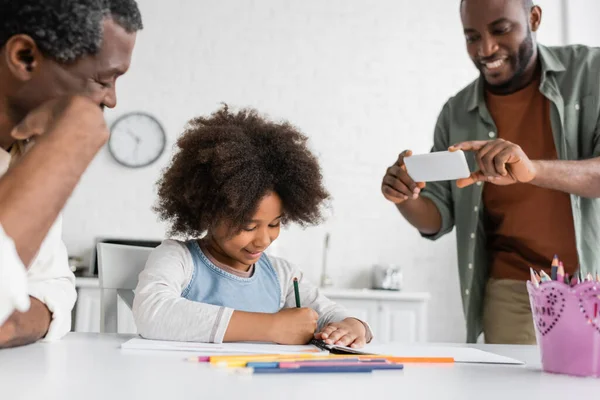 Hombre afroamericano feliz tomando fotos en el teléfono inteligente mientras su hija y su padre dibujan juntos - foto de stock