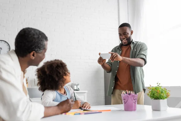 Cheerful african american man taking photo on smartphone while his daughter and father drawing together — Stock Photo