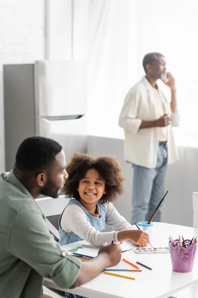 Smiling african american girl painting near father and blurred granddad at home — Stock Photo