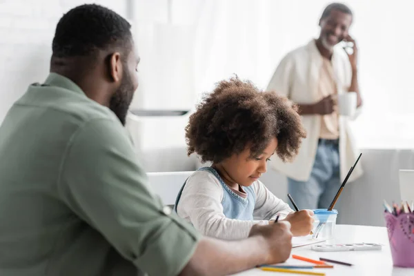 Chica afroamericana pintando en cuaderno de bocetos cerca de padre borroso y abuelo en casa - foto de stock