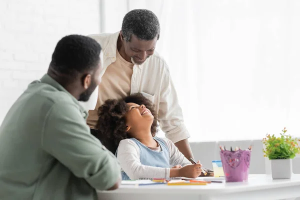 Cheerful african american girl looking at granddad while painting with father at home — Stock Photo