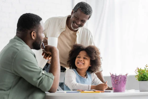 Positive african american child painting and looking at blurred dad near grandfather with coffee at home — Stock Photo