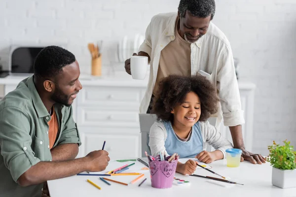 Positive african american man sitting near daughter painting and father holding cup of coffee at home — Stock Photo