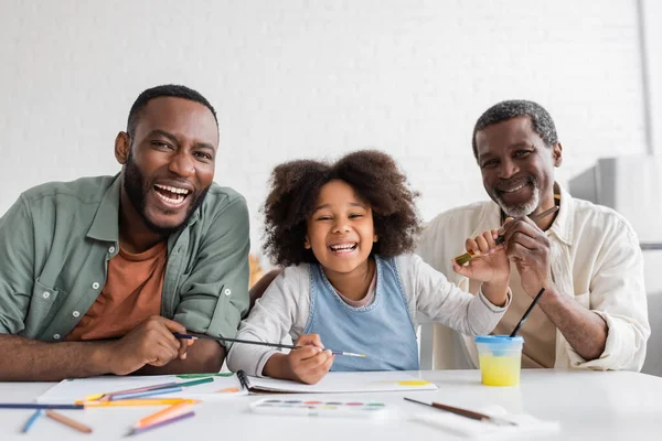 Smiling african american girl looking at camera near father and granddad while painting at home — Stock Photo