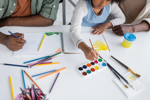Vista recortada de niños afroamericanos y padres pintando en cuaderno de bocetos en casa - foto de stock