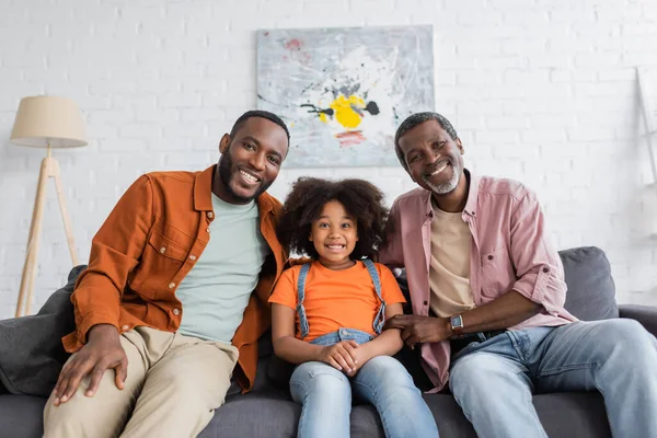 Sonriente chica afroamericana mirando a la cámara cerca de papá y abuelo en el sofá en casa - foto de stock