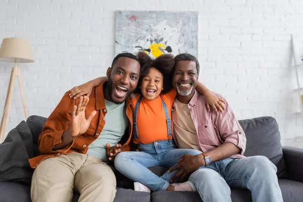 Alegre afroamericano chica abrazando abuelo y papá en sofá en sala de estar - foto de stock