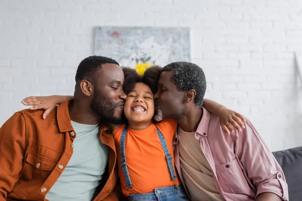 Padre y abuelo afroamericanos besando a un niño alegre en la sala de estar - foto de stock