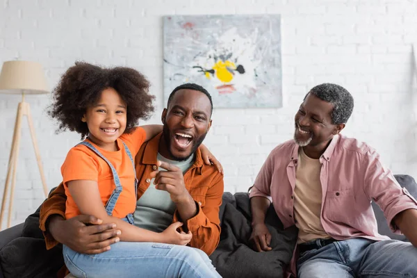 Positive african american man pointing with finger and holding daughter near grandfather on couch at home — Stock Photo