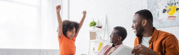 Garota americana africana excitada mostrando sim gesto perto dos pais com jornal em casa, banner — Fotografia de Stock